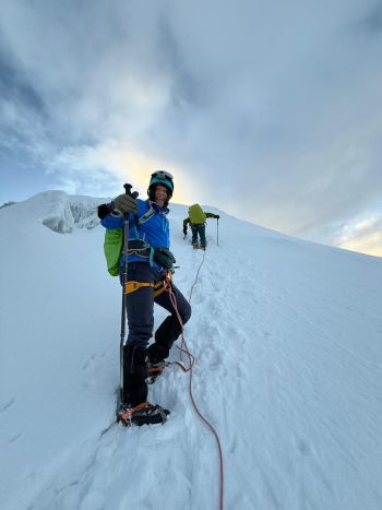 A smiling mountaineer in blue jacket and helmet gives a thumbs-up while roped to a guide climbing the icy slopes of Mount Chimborazo, Ecuador.