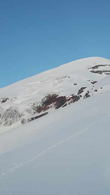 A climber's boot trail cuts across a smooth snowfield leading toward exposed red volcanic rock and the snow-capped peak of Mount Chimborazo in Ecuador.