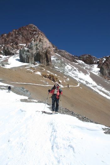 A mountaineer with trekking poles traverses a high-altitude snow trail on Mount Aconcagua beneath rugged rock formations and a clear blue sky in Mendoza, Argentina.