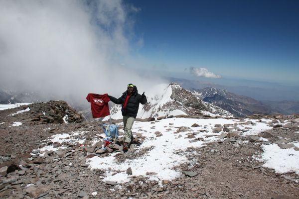 A climber proudly holds a t-shirt and gives a thumbs-up at the summit of Mount Aconcagua, surrounded by snowy ridges and dramatic clouds in Mendoza, Argentina.