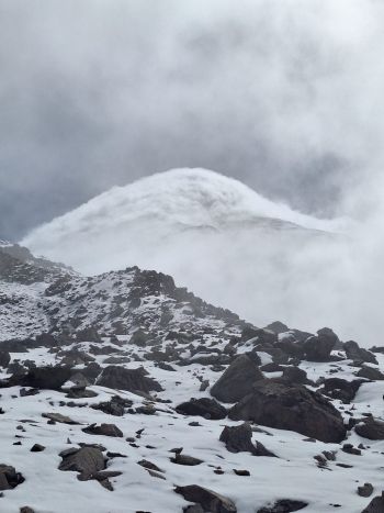 Dark volcanic rocks and snowpatches line the foreground with Mount Chimborazo's summit wrapped in dense, dramatic clouds in Ecuador.