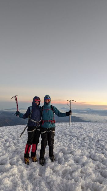 Two mountaineers hold ice axes high in celebration on the snowy summit of Mount Chimborazo, Ecuador, as the sun rises over distant cloud horizons.