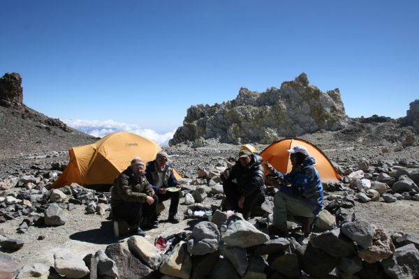 A team of mountaineers sharing a meal outside their tents at a high-altitude camp on Mount Aconcagua, surrounded by rocky terrain and clear skies in Mendoza, Argentina.