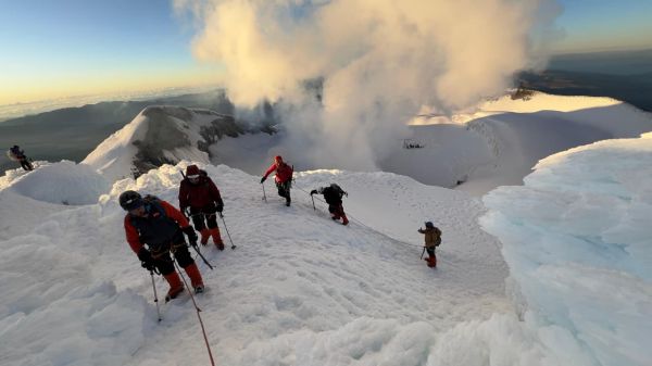 Roped mountaineering teams navigate icy snow ridges near the summit of Mount Chimborazo during sunrise, with dramatic steam clouds in Ecuador.