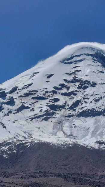 A telephoto shot capturing the snow-dusted summit dome and icy rock walls of Mount Chimborazo under a clear blue sky in Ecuador.