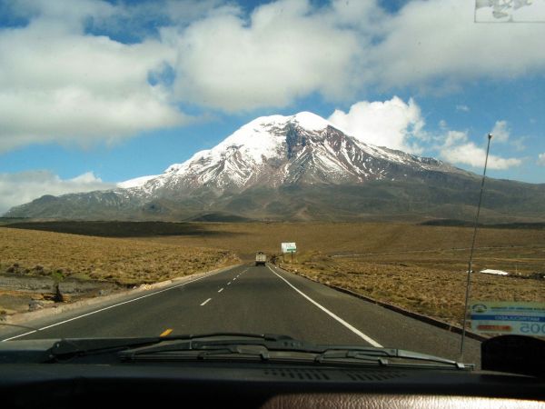 A driver's perspective along a paved highway heading toward the massive snow-capped Mount Chimborazo volcano in Ecuador.