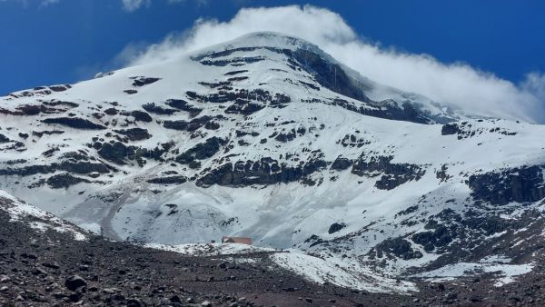 A mountain refuge building sits beneath the massive, snow-dusted peak and rocky glaciers of Mount Chimborazo under a blue sky in Ecuador.