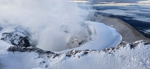 A dramatic panoramic view looking down into the snow-dusted, steaming active crater from the summit of Cotopaxi Volcano in Ecuador.
