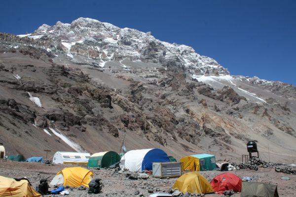 A vibrant high-altitude base camp featuring colorful expedition tents set against the rugged, snow-capped slopes of Mount Aconcagua in Mendoza, Argentina.