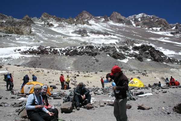 A group of mountaineers resting at a high camp on Mount Aconcagua, Mendoza, surrounded by rocky, snow-dusted Andean peaks under a clear sky.