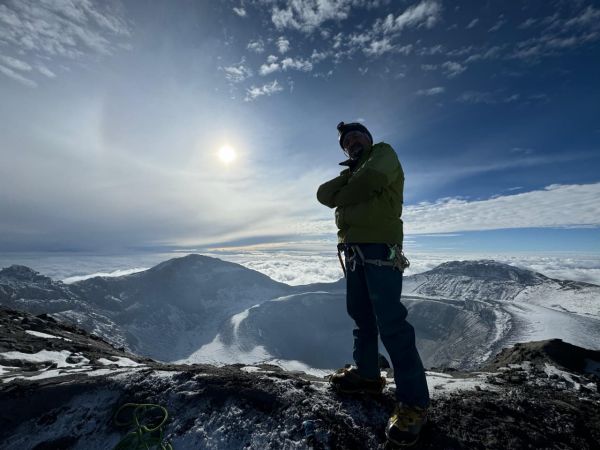 A climber stands proudly at the summit rim of Cotopaxi Volcano in Ecuador under a bright sun, overlooking the massive snow-dusted crater and cloud layer.