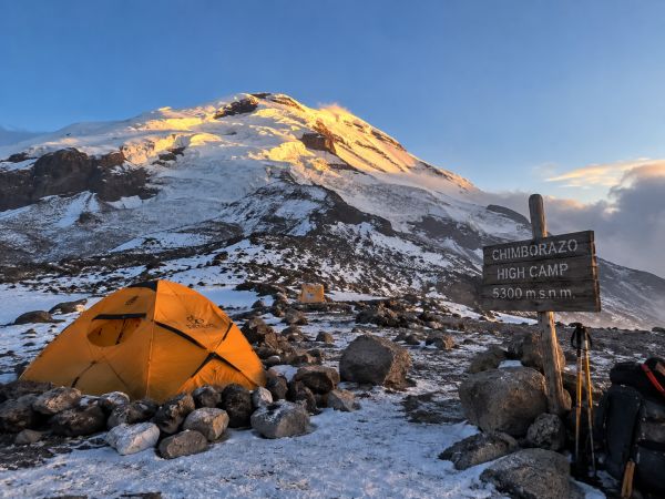 A high-altitude expedition tent sits at Chimborazo High Camp (5,300m / 17,388ft) beside a wooden trail sign as golden hour sunset light strikes the volcano peak in Ecuador.